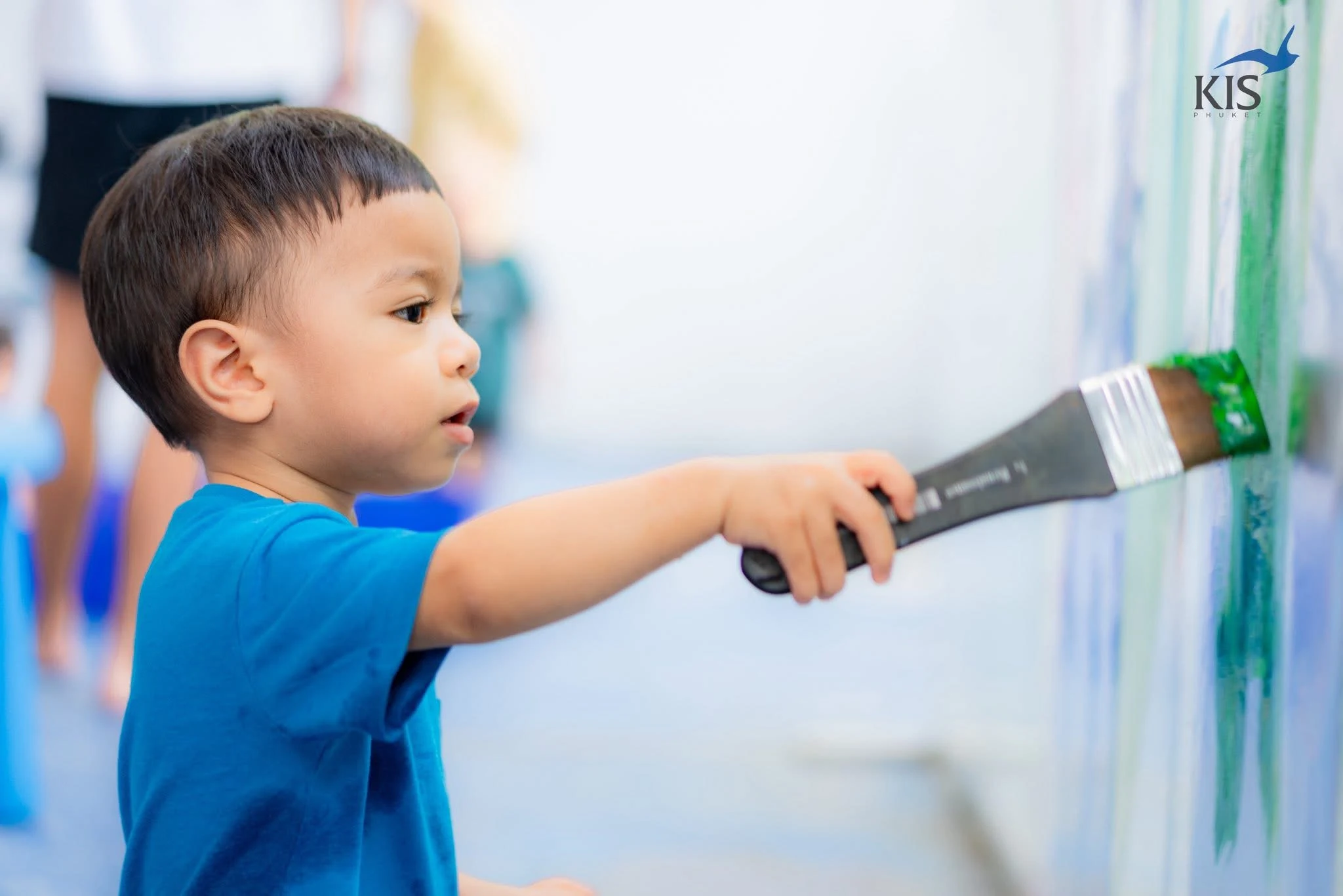 A child in a blue shirt painting with a large brush in a bright activity room