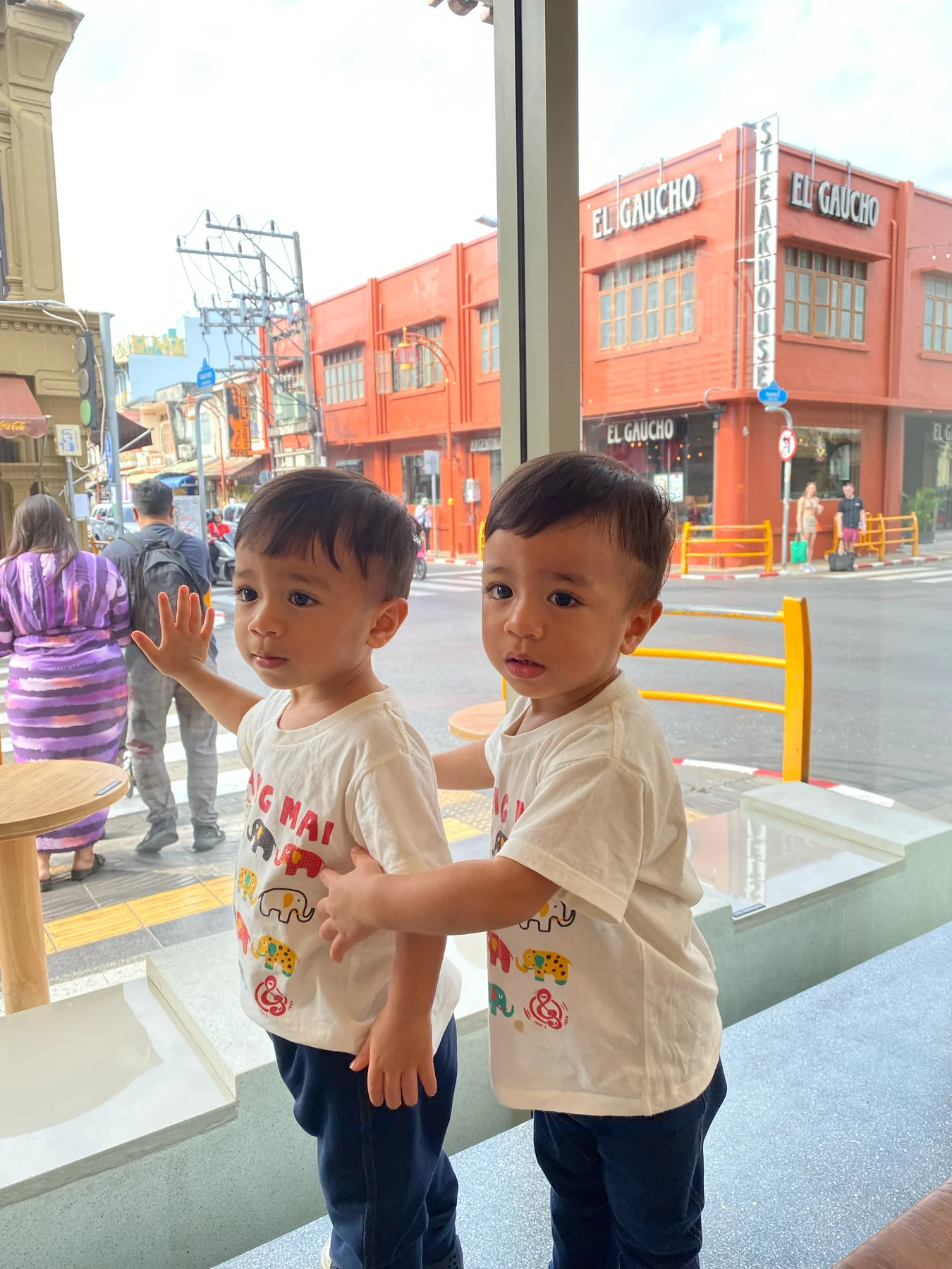 Two children standing by a cafe window overlooking a colorful street