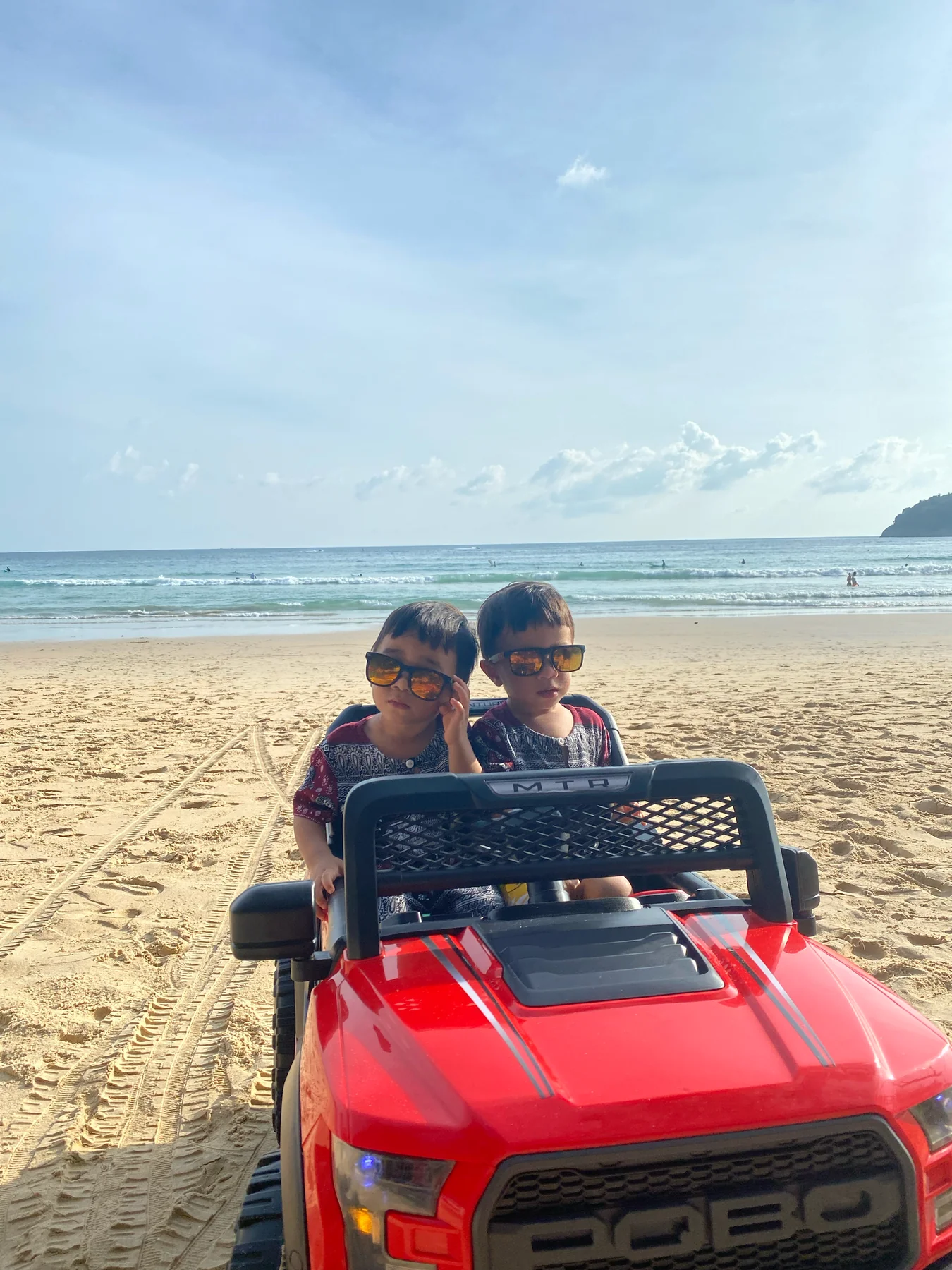 Two children wearing sunglasses in a red toy car by the sea