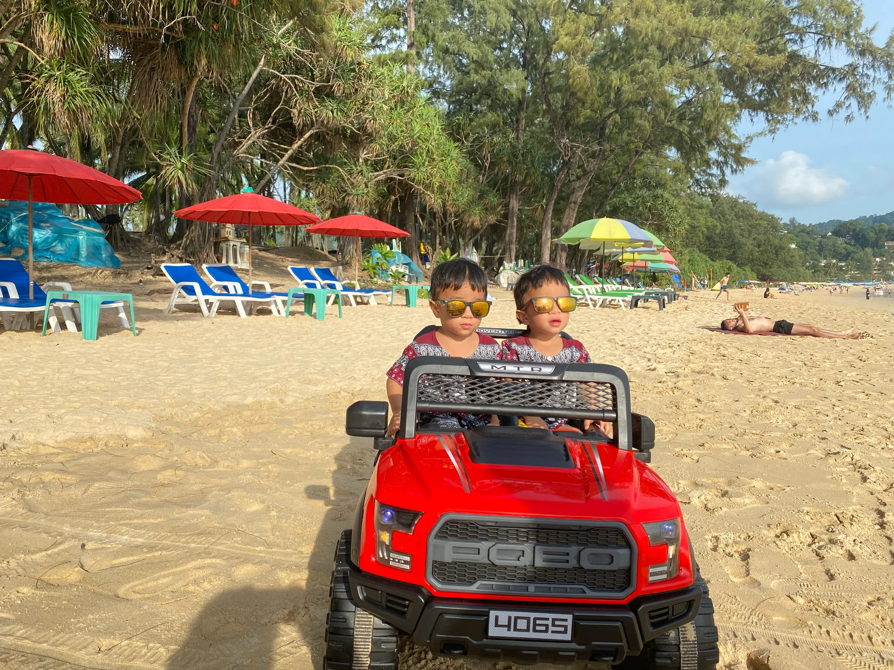 Two children in a red toy car on a beach with colorful umbrellas